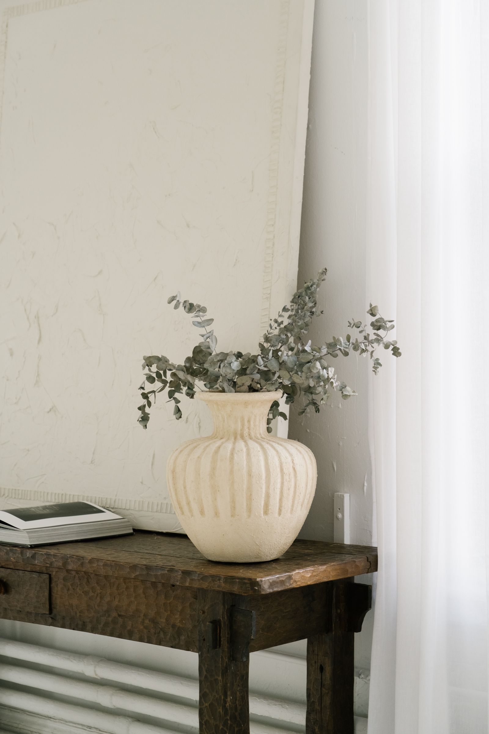 A ceramic vase of dried branches on a wooden table beside a sheer curtain.