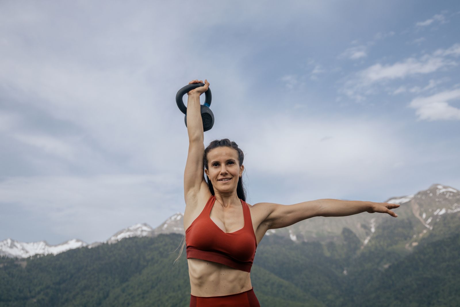 A strong, smiling woman lifts a kettlebell overhead on a hillside with mountains behind her.