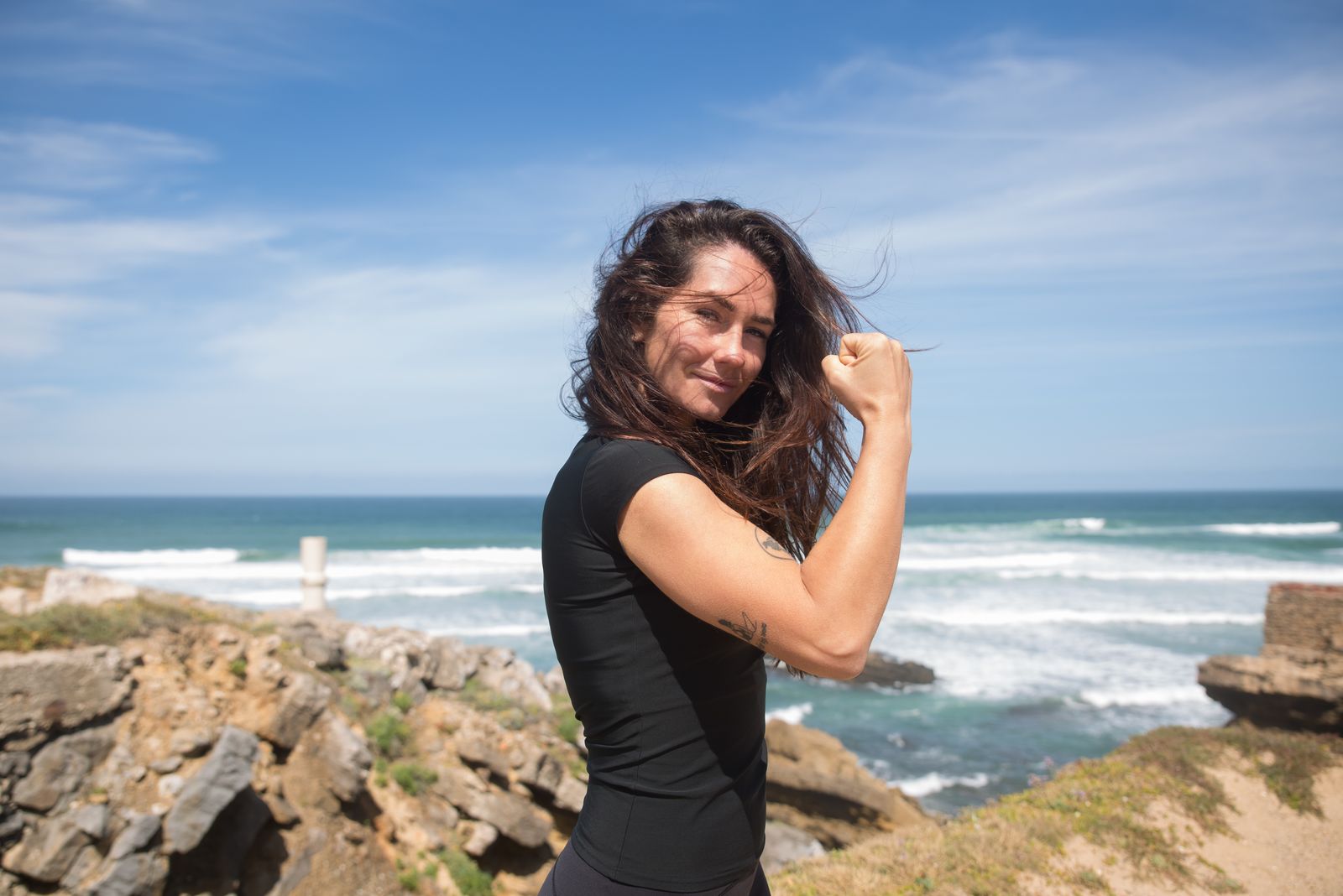 A confident woman flexes her arm by the sea, her hair blowing in the wind.