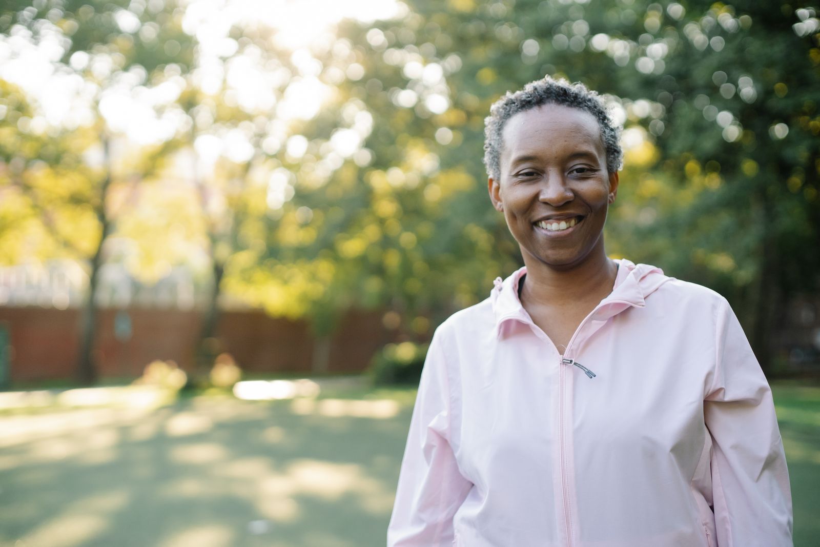 A smiling woman with short grey-streaked hair in a soft pink jacket stands in a sunny garden.